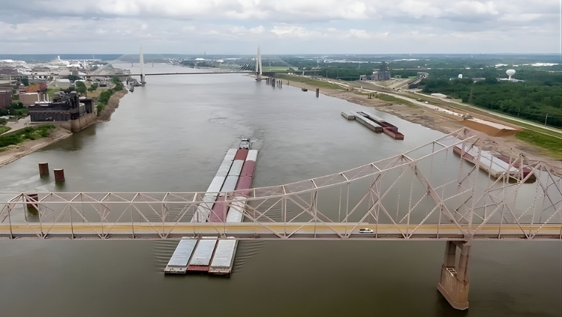 Barges transport cargo along the Mississippi River beneath steel bridge near industrial port facilities